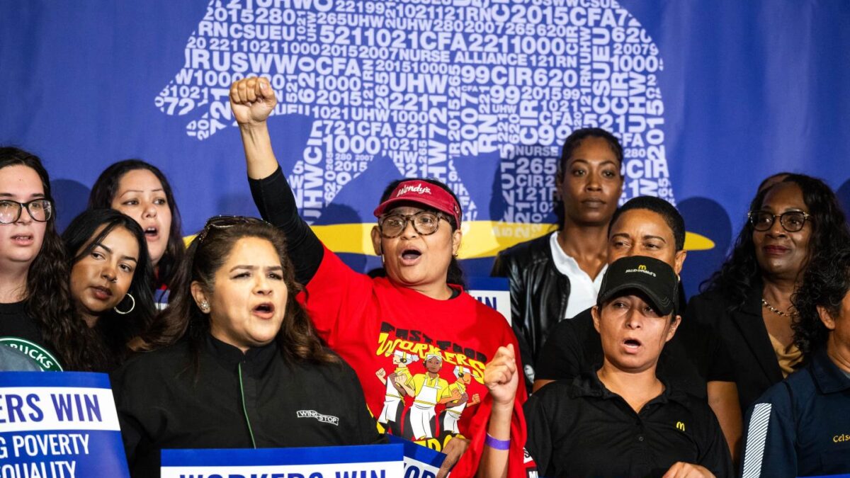 A woman in a red shirt, surrounded by other people, cheers and holds a fist in the air.