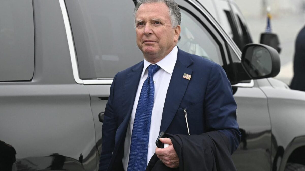 Steve Witkoff walks in front of a car dressed in suit and tie with his phone in his hand and his jacket draped over his arm.