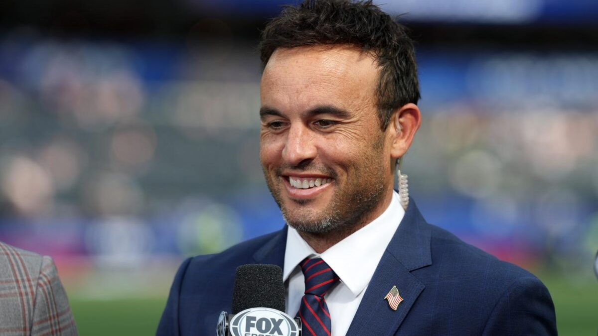 Landon Donovan, wearing a suit and holding a microphone, smiles.